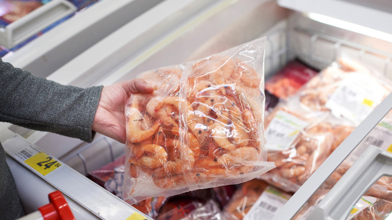 A hand holding a bag of shrimp over a grocery store freezer.