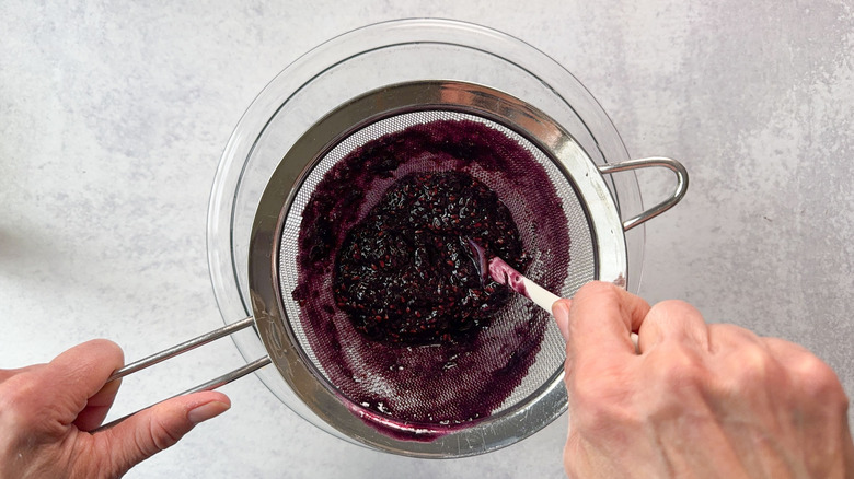 Pushing blackberry puree through fine mesh strainer over glass bowl with spatula
