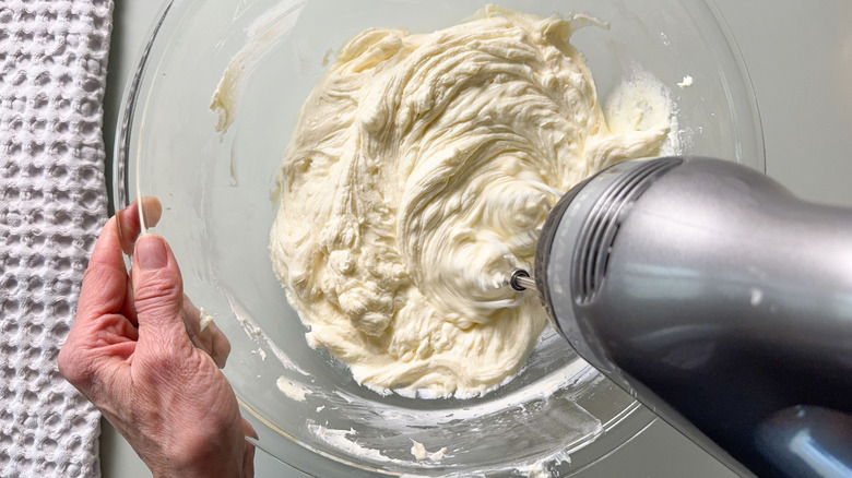 Beating cream cheese, sour cream, and sugar in glass mixing bowl with hand mixer