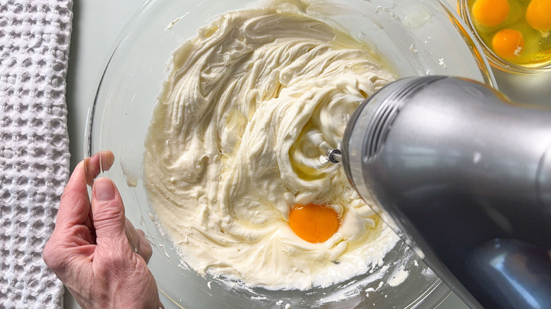 Adding eggs to cheesecake batter with hand mixer in glass bowl