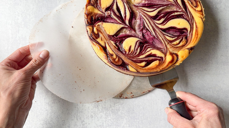 Lifting blackberry marbled cheesecake off parchment paper circle with spatula