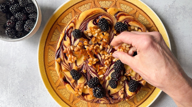Garnishing marbled blackberry cheesecake with candied pine nuts on platter next to bowl of fresh blackberries