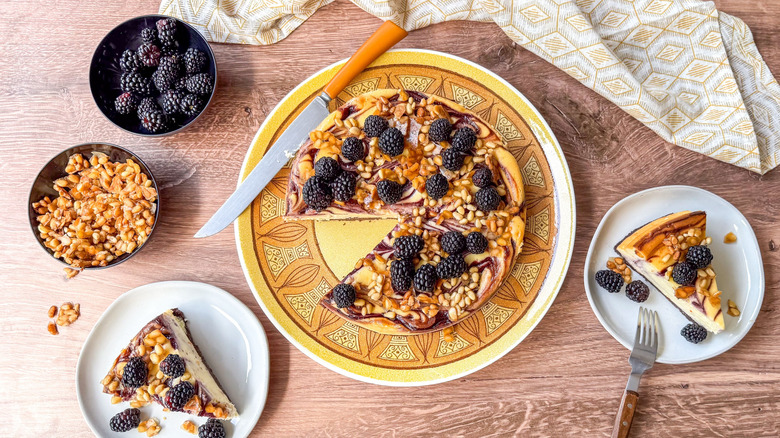 Blackberry and pine nut cheesecake on serving platter with slices on plates and bowls of candied pine nuts and fresh blackberries