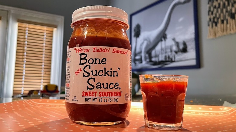 A jar of Bone Suckin' Sauce sits on a table next to a glass filled with sauce.