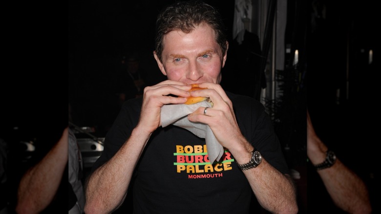 Bobby Flay in black Bobby's Burgers T shirt eating a burger