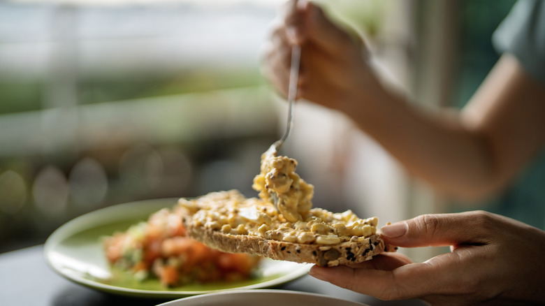 A person's hand is seen putting egg salad on bread with a spoon.
