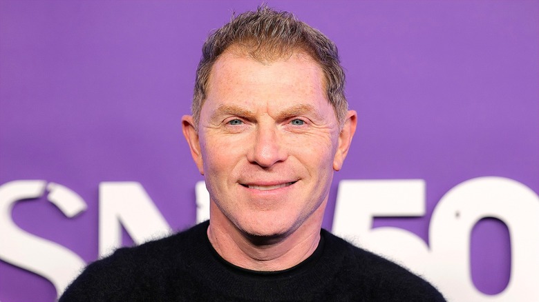 Bobby Flay smiling in a black sweater in front of a purple backdrop with white text