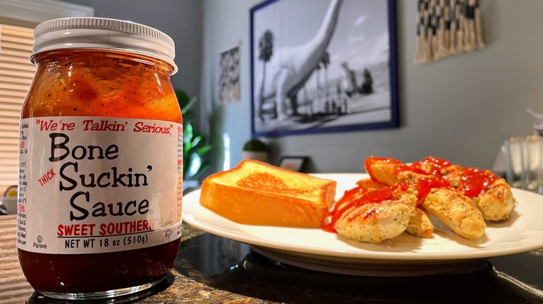 A jar of Bone Suckin' Sauce sits next to a plate with grilled chicken that has sauce on it.