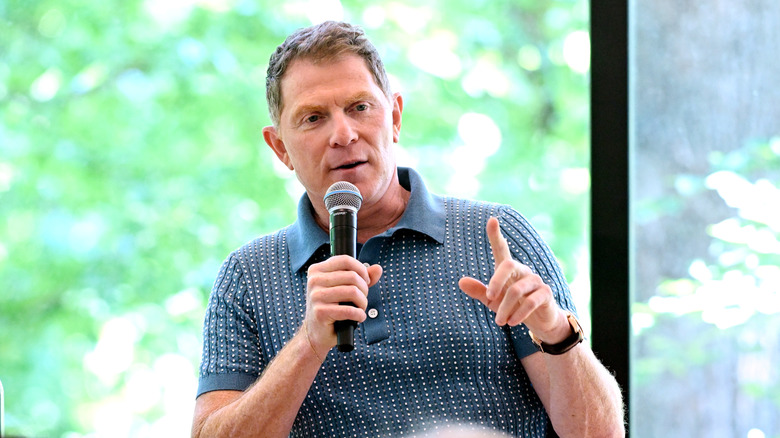Celebrity chef Bobby Flay sits on stage at an event pointing with one hand and holding a microphone with the other