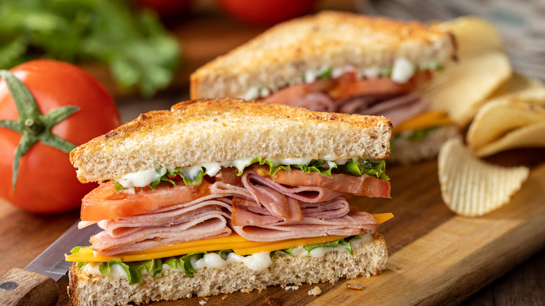 Toasted deli sandwich on wooden cutting board with fresh tomato and potato chips.