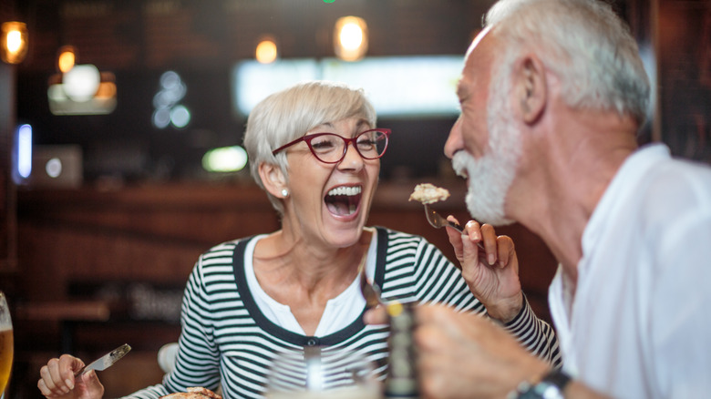 Older woman smiling while feeding an older man a bite of food in a restaurant