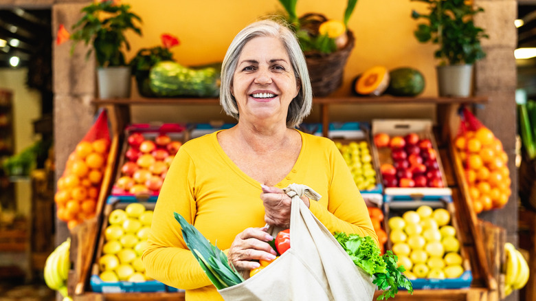 Senior woman in a yellow shirt smiling and holding a reusable bag full of groceries in front of a produce display