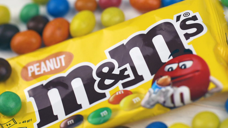 A closeup of a bag of peanut M&M's with loose candies in the background.