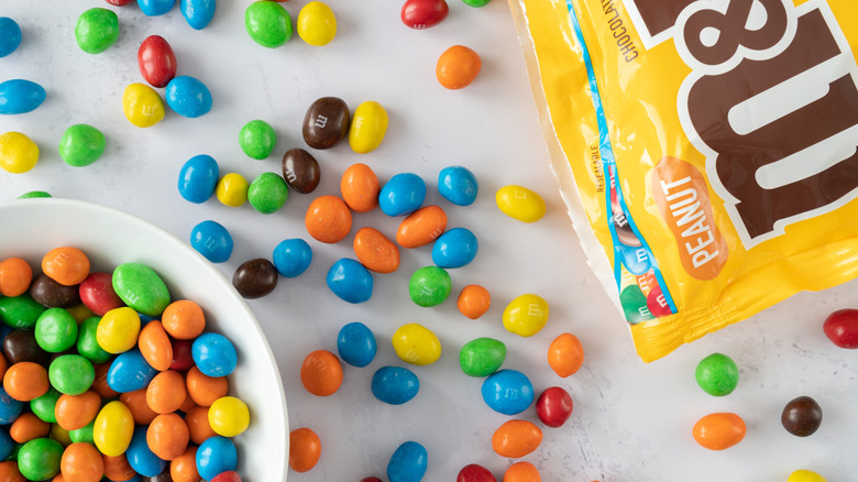 An open bag of peanut M&Ms with candies in a bowl and scattered on a tabletop