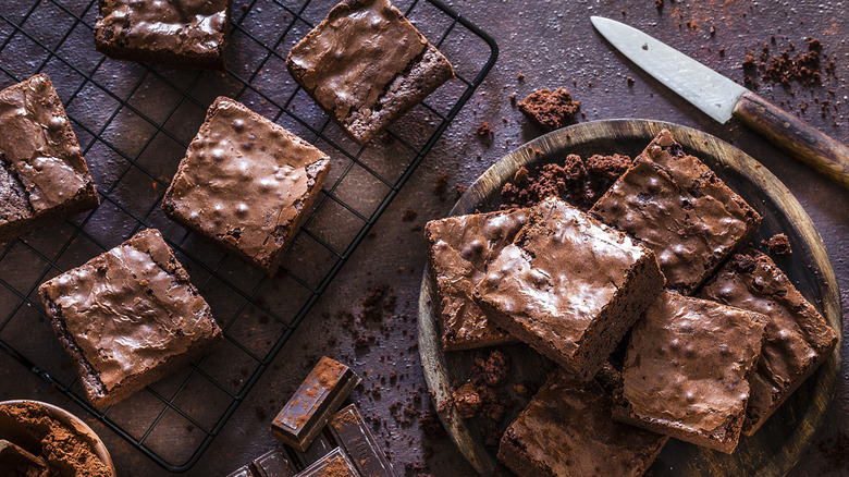 A plate of brownies next to a wire rack of brownies with crumbs scattered throughout the background