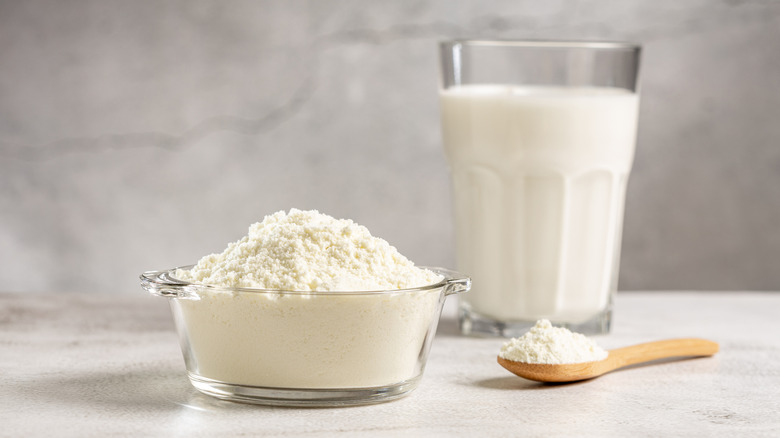 A small bowl of powdered milk next to a spoonful of powdered milk and in front of a glass of liquid milk.