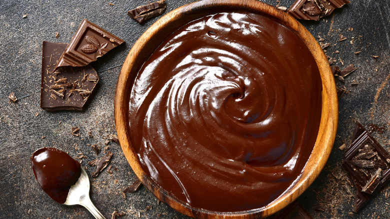 Bowl of shiny chocolate ganache on gray slate with pieces of broken chocolate