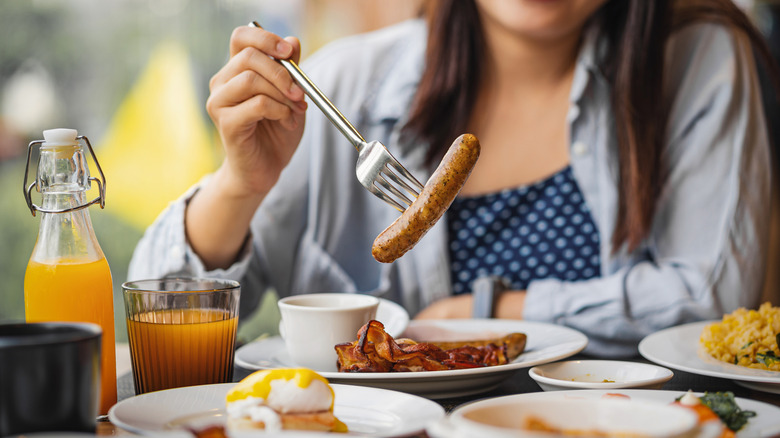 A person's hand spears a big breakfast sausage link with a fork at a bountiful breakfast table