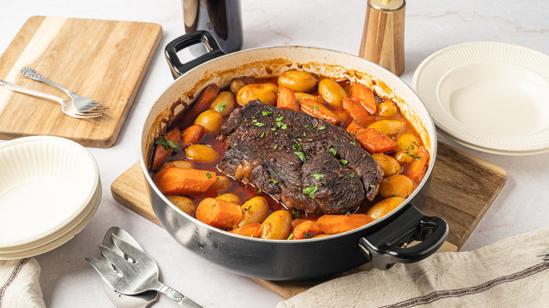 Pot roast is surrounded by carrots and potatoes in a round pan on a cutting board next to bowls and serving utensils