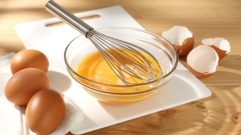 Beaten eggs in bowl with whisk on white cutting board with whole eggs on the side.