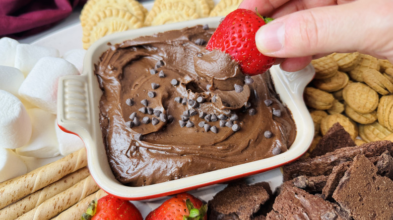 Hand dipping strawberry into brownie batter dip surrounded by other dippers