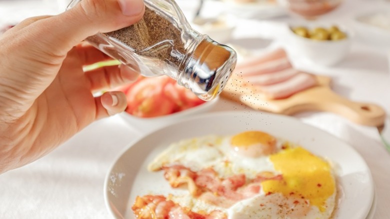 A hand seasoning a plate of bacon and eggs