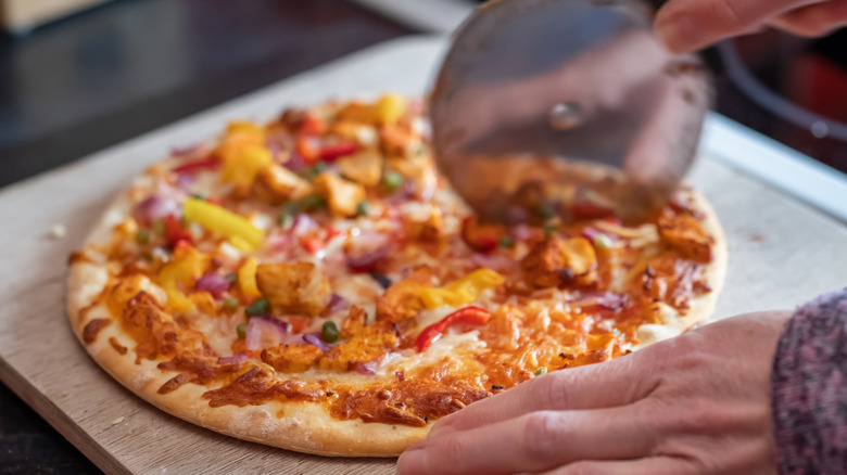 Close-up of hand cutting baked frozen pizza with a pizza cutter on a wooden board.