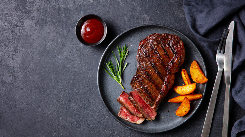A plated cooked steak with potatoes and a sprig of fresh rosemary, next to silverware and a ramekin of sauce