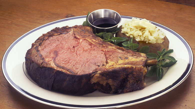 Large slice of prime rib on a white plate with herbs, a baked potato, and a side of au jus