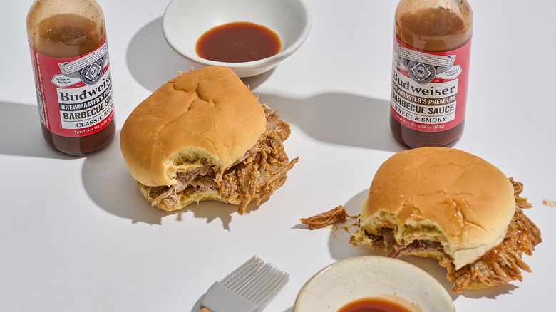 Pulled pork sandwiches on a table with barbecue sauce bottles and bowls of barbecue sauce