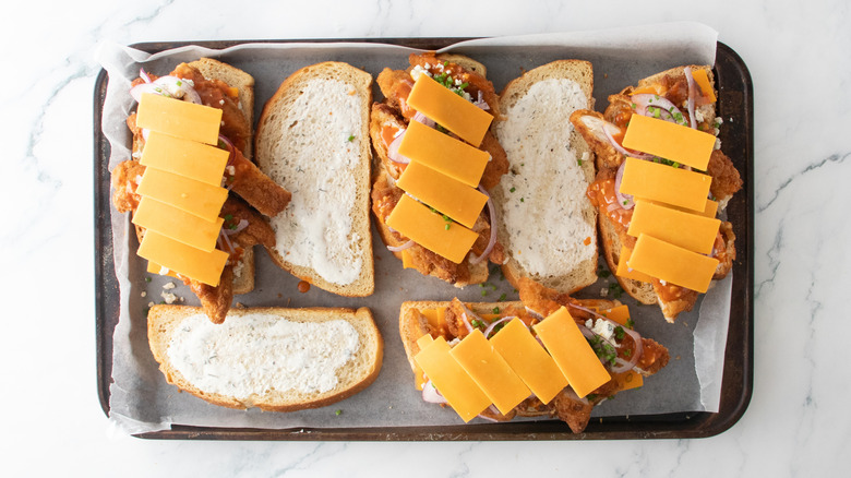 A large baking sheet containing multiple bread slices topped with chicken tenders and cheeses.