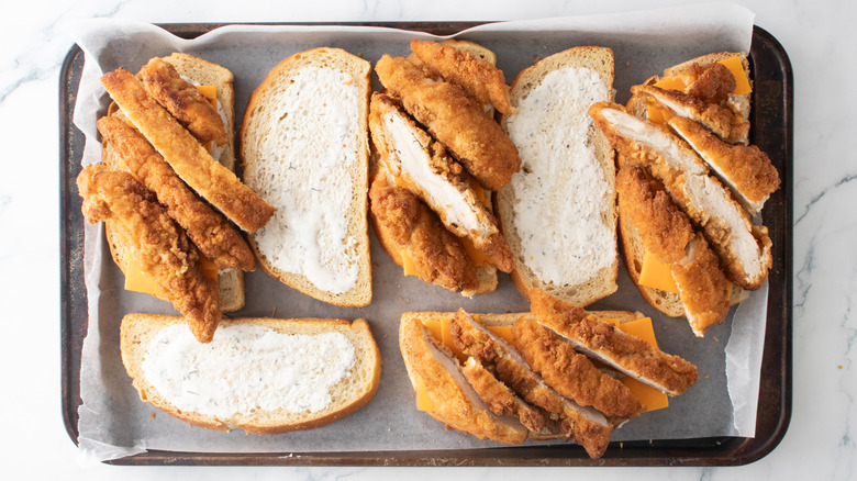 A large baking sheet with multiple bread slices containing slices of cheddar cheese and sliced chicken tenders.