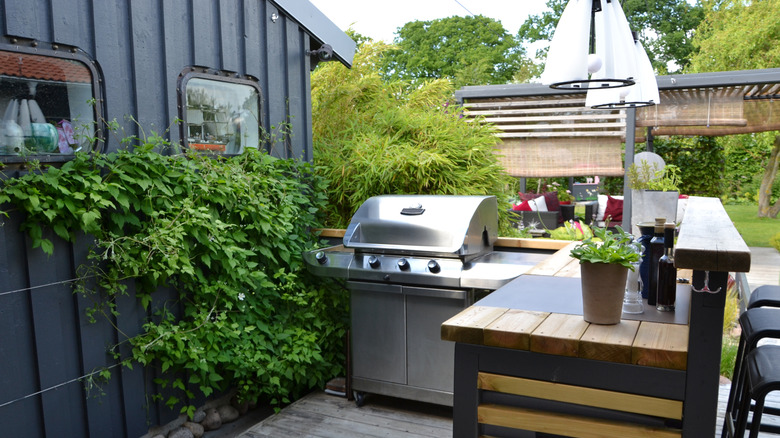 A grill and wooden countertop are nestled against the wall of a house with lots of plants