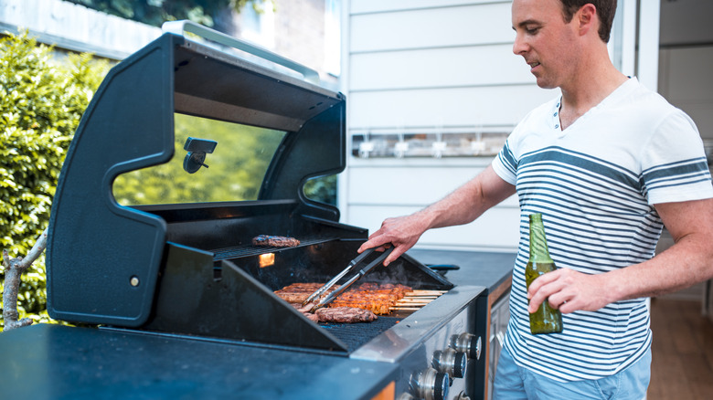 A person grills burgers and sausages in an outdoor kitchen while drinking a beer