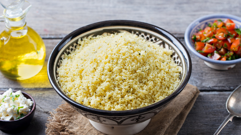 A bowl of fluffy couscous surrounded by fresh ingredients, including diced tomatoes, olive oil, and feta cheese