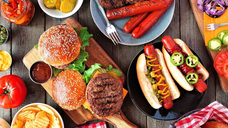 Grilled burgers and hot dogs surrounded by various toppings like sliced cheese, red onion slices, tomatoes, and pickles