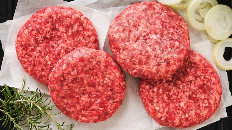 Four ground beef patties on white squares of parchment paper surrounded by fresh rosemary and sliced onions