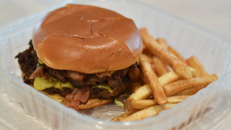 A Chili's bacon burger in a clear container with fries on a white countertop