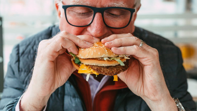 Older man biting into a burger, squeezing toppings out