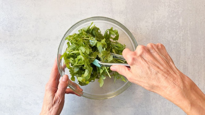 Tossing fresh arugula with olive oil using tongs in glass bowl