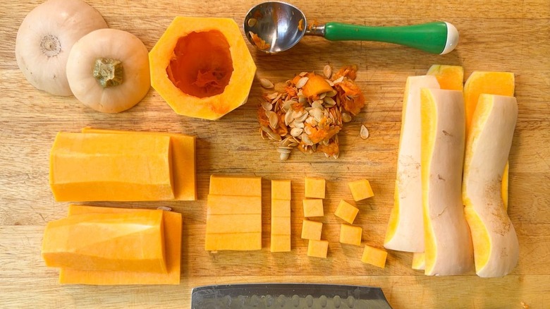 Butternut squash peels, seeds and flesh cut into sections and cubes on cutting board with ice cream scoop