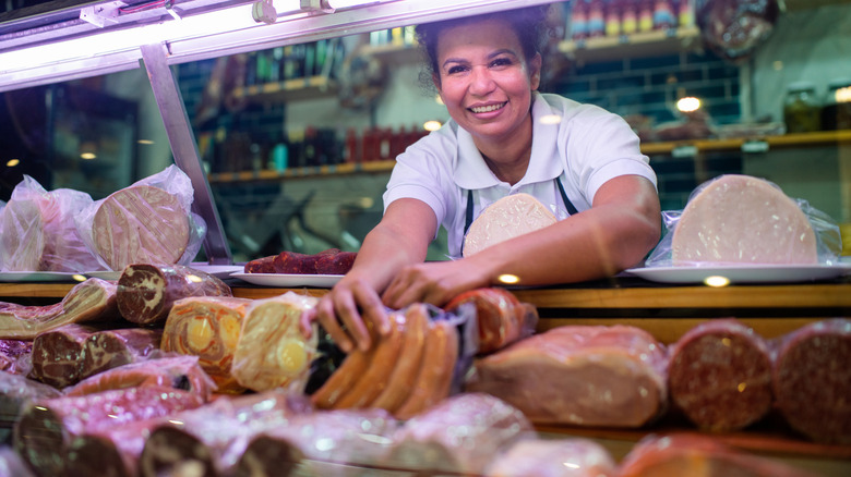 A woman smiling at a meat counter holding up a package of hotdogs.