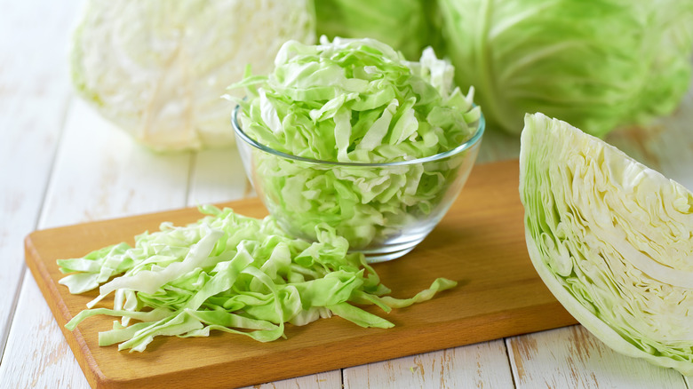 A bowl of chopped up cabbage on a cutting board, surrounded by other cabbages