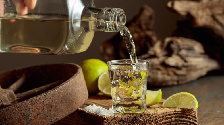 Tequila being poured into a shot glass surrounded by lime slices and salt.