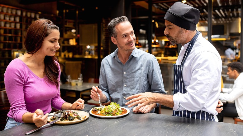 A chef talking to a man and woman while they enjoy two separate meals