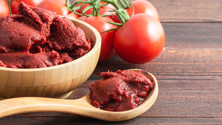 A small wood bowl of tomato paste next to a wood spoon with tomato paste and fresh tomatoes in the background on wood surface