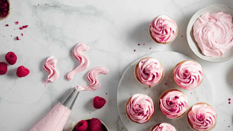 Pink cupcakes with bowl of pink frosting, a piping bag, and raspberries
