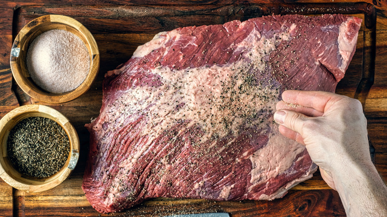 Raw brisket being seasoned on a wooden cutting board.