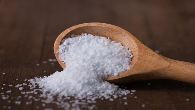 Wooden spoon with salt flakes spilling from it onto wood surface.
