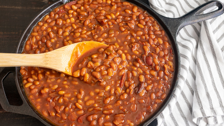 Baked beans being stirred with a wooden spoon in a cast iron skillet